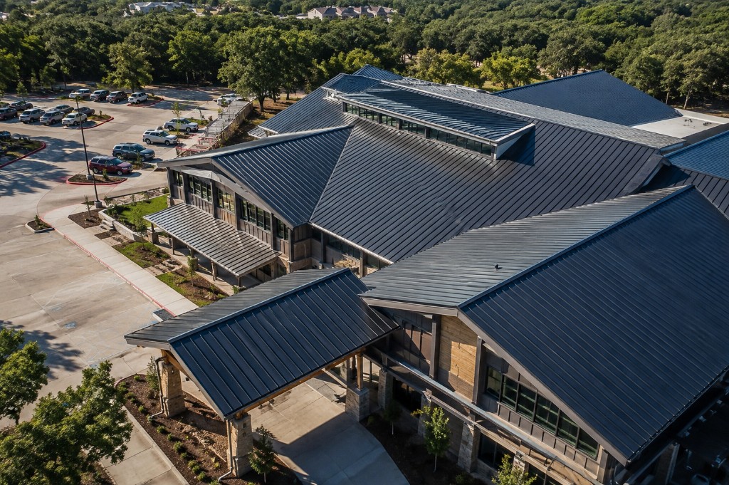 Aerial view of a commercial building with a standing seam metal roof and building envelope, surrounded by parking and mature trees